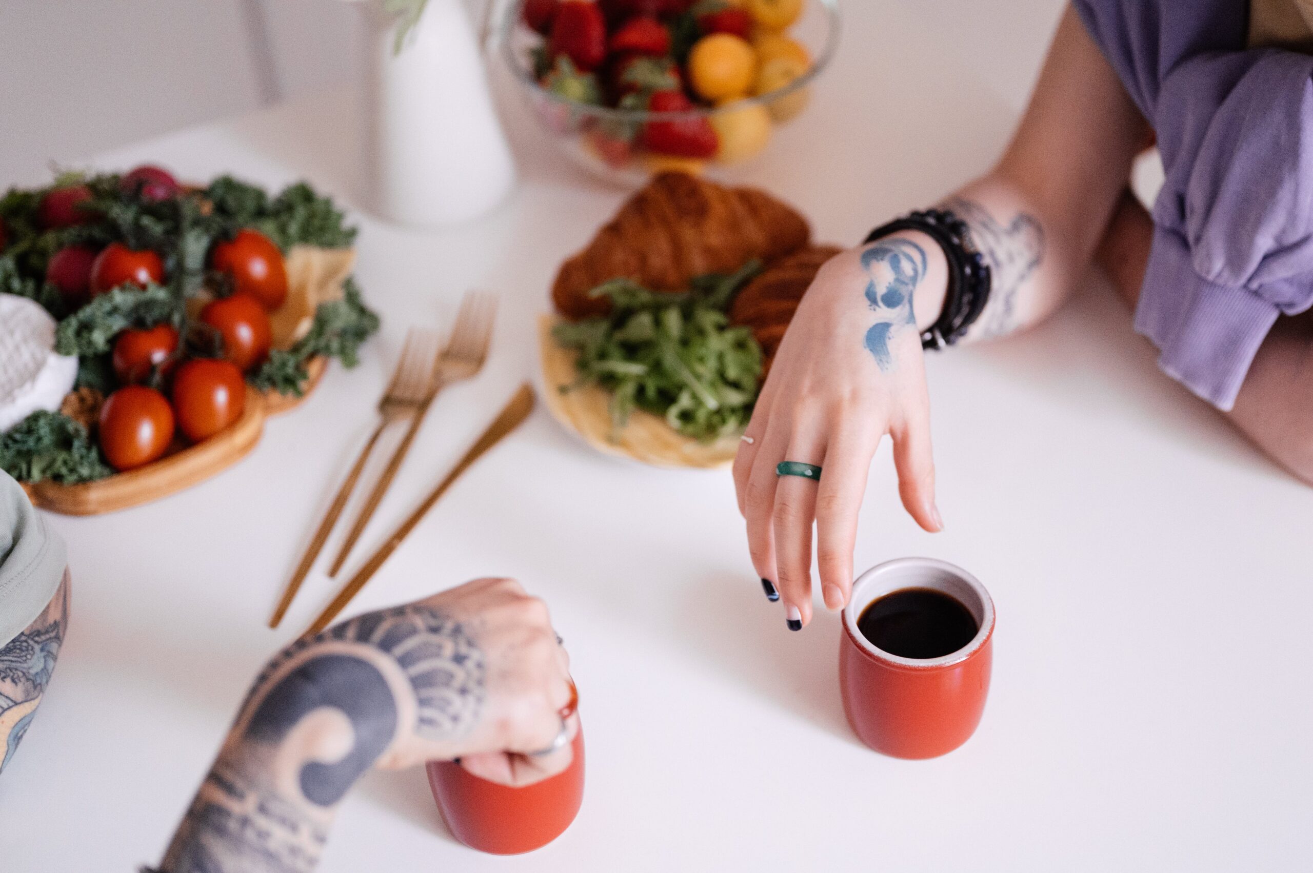 Hands of two people with tattoos o their hands and forearms, sharing a cup of coffee at a table with plates and bowls of food. Both hands have rings on them. The hand facing the camera belongs to a person with black and white painted fingernails, a black bracelet, and a lavender blouse. Their faces cannot be seen.